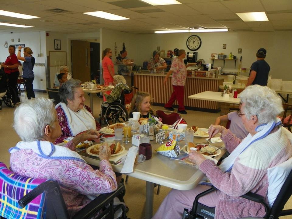 People dining at a communal table in what appears to be a care facility, with staff in the background.