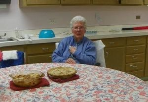 An elderly woman sitting at a table with two pies in a kitchen with vintage decor.