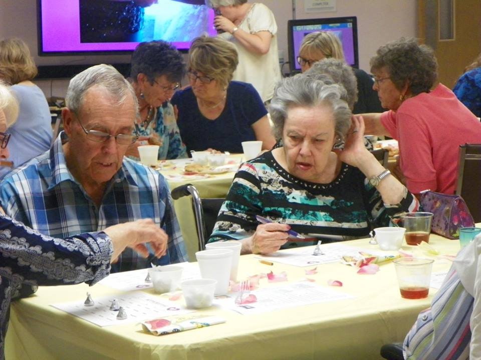A group of people, possibly seniors, engaged in an activity around tables with papers and small cups.