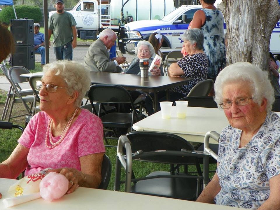 An outdoor gathering with older adults sitting at tables, some holding drinks, with a police car and speaker in the background.