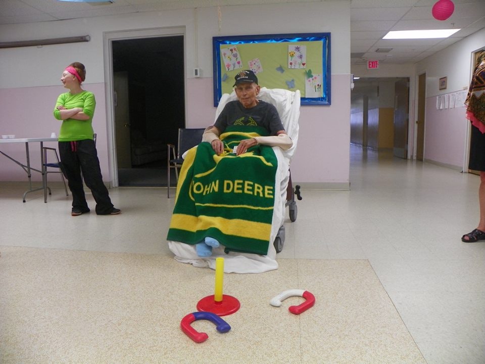An indoor gathering with a person in a wheelchair holding a blanket, horseshoe game pieces on the floor, and two standing people.
