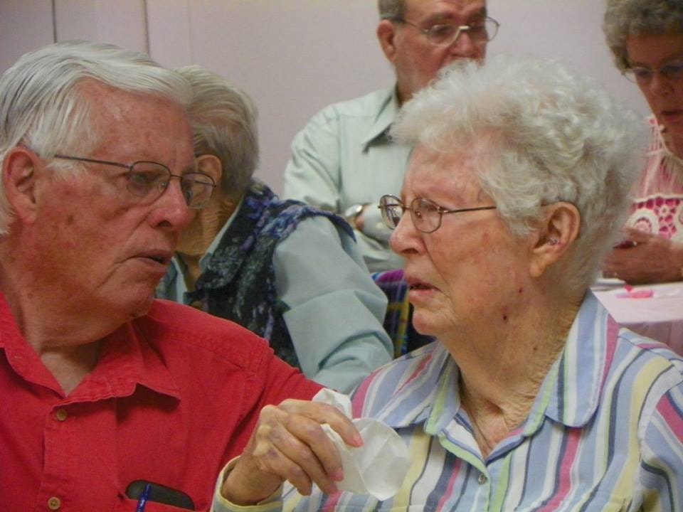 An elderly couple converses at a social event, surrounded by others in a room with a pink wall.