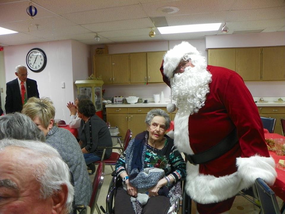 Person dressed as Santa Claus interacting with elderly individuals in an indoor setting, festive decorations visible.