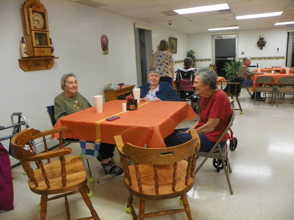 Three people seated around a table with orange tablecloth in a room with others and wall decorations.