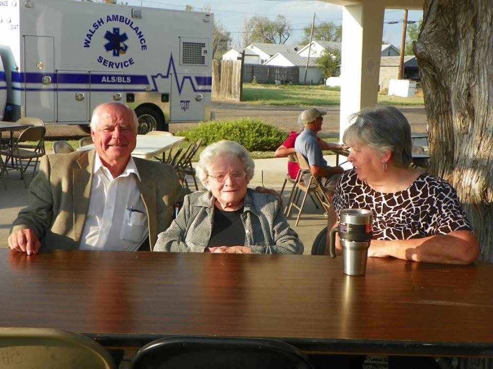 Three people sitting at a table outdoors with an ambulance in the background.