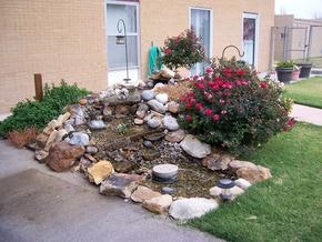A garden with rocks, greenery, and red flowers beside a building.