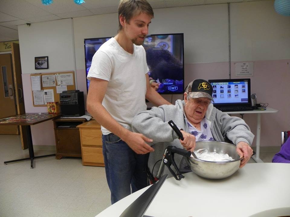 A younger man assists an elderly person with cooking in a kitchen setting.
