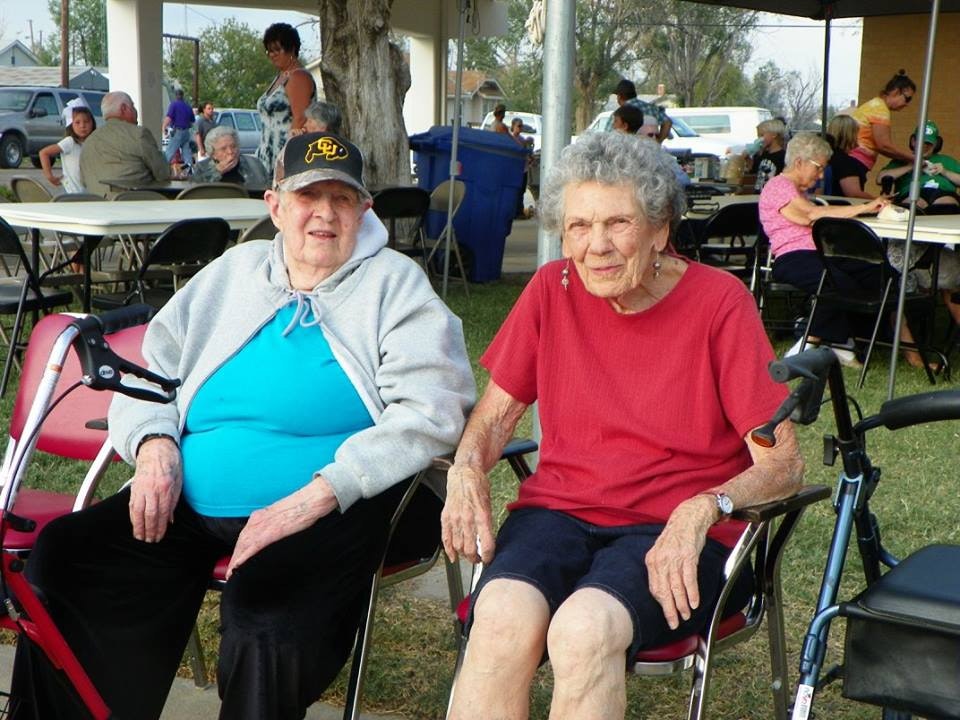 Two elderly individuals in a park with others, some seated, and walking aids nearby.