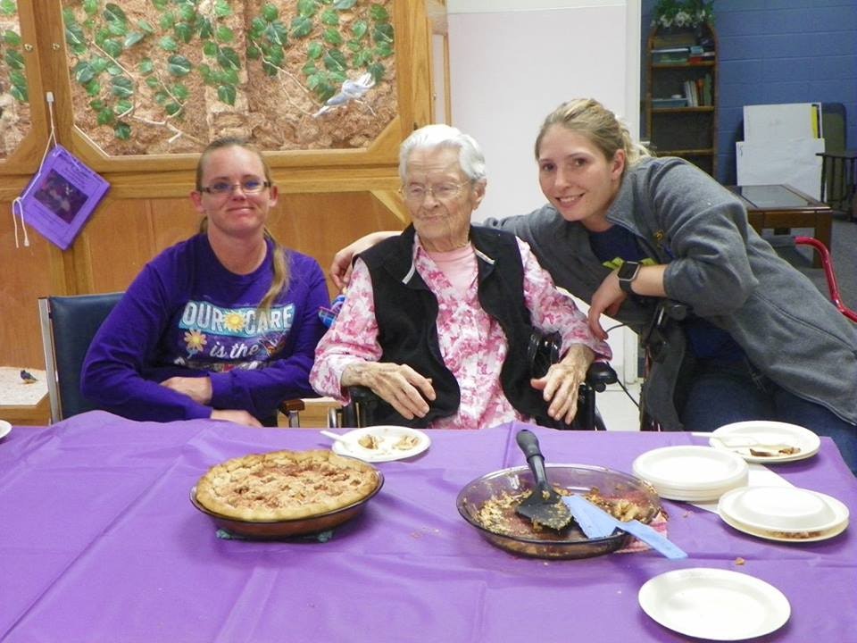 Three people smiling at a table with a pie and plates in a room with a purple tablecloth.