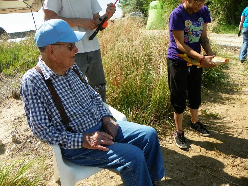 Elderly man seated outdoors, people standing nearby, sunny day, casual setting, someone holding a tool or device.