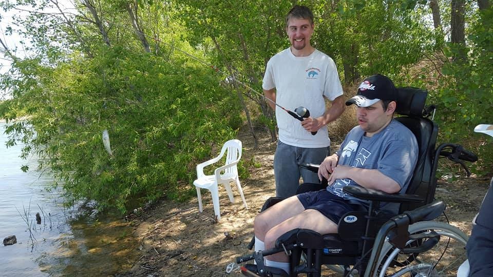 Two people by a lake, one standing with a fish on a line, the other in a wheelchair, trees and chairs around.
