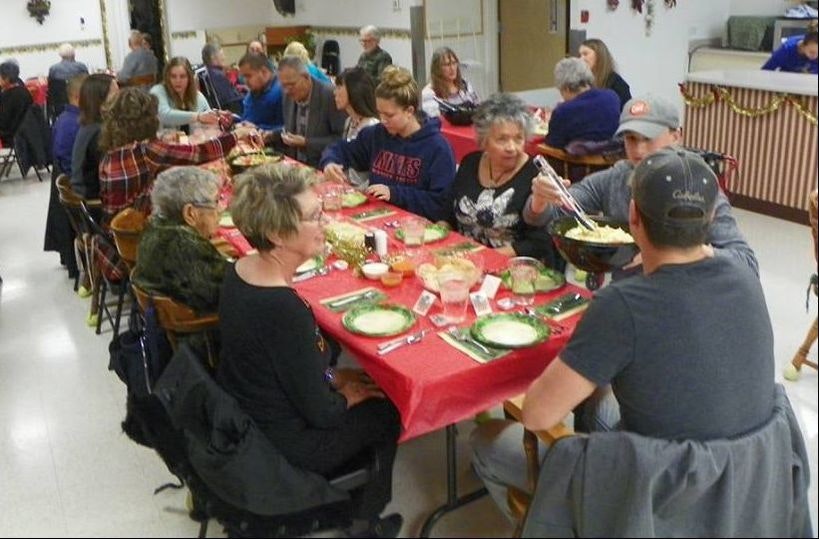 Group of people dining at a long table with red tablecloth, plates, and food. Social gathering indoors.