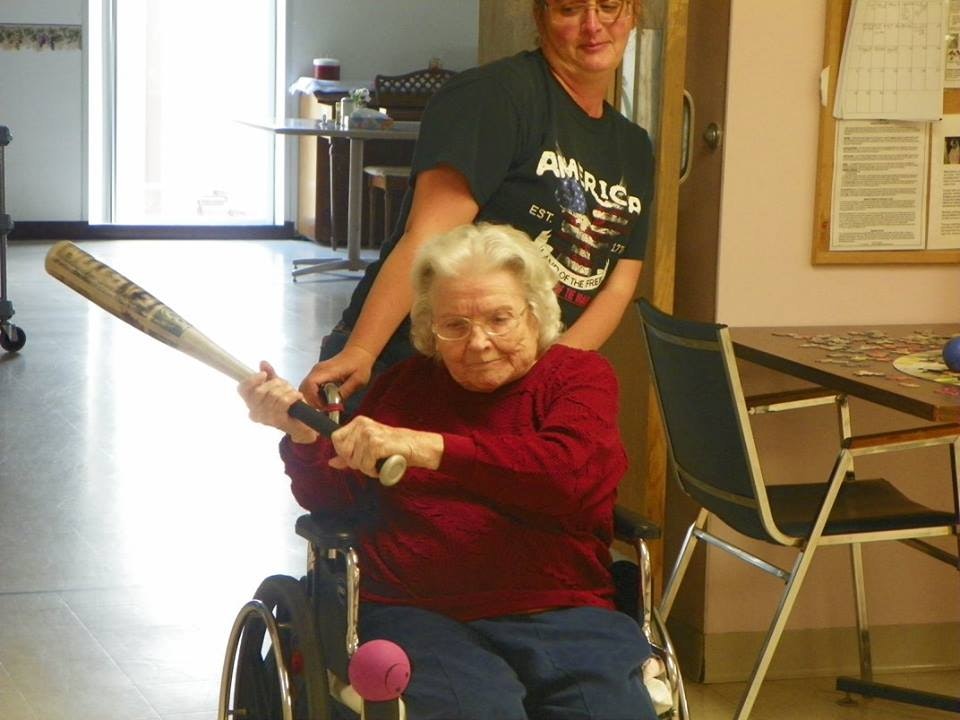Elderly woman in a wheelchair playing baseball indoors with assistance.