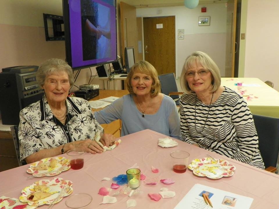 Three women smiling at a table with snacks, drinks, and a TV in the background. Looks like a social gathering.