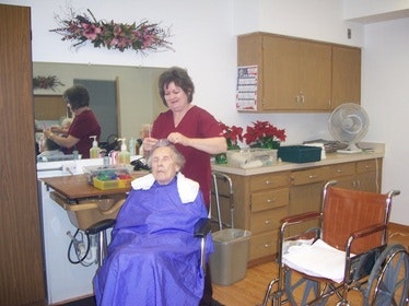 A person in a salon cape sits in a wheelchair while another person styles their hair, in a salon environment.