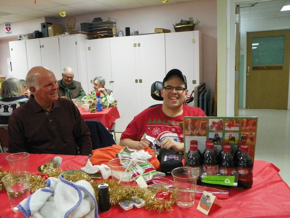 People at a festive gathering with holiday decorations, gifts, and a man smiling with beverages in front of him.