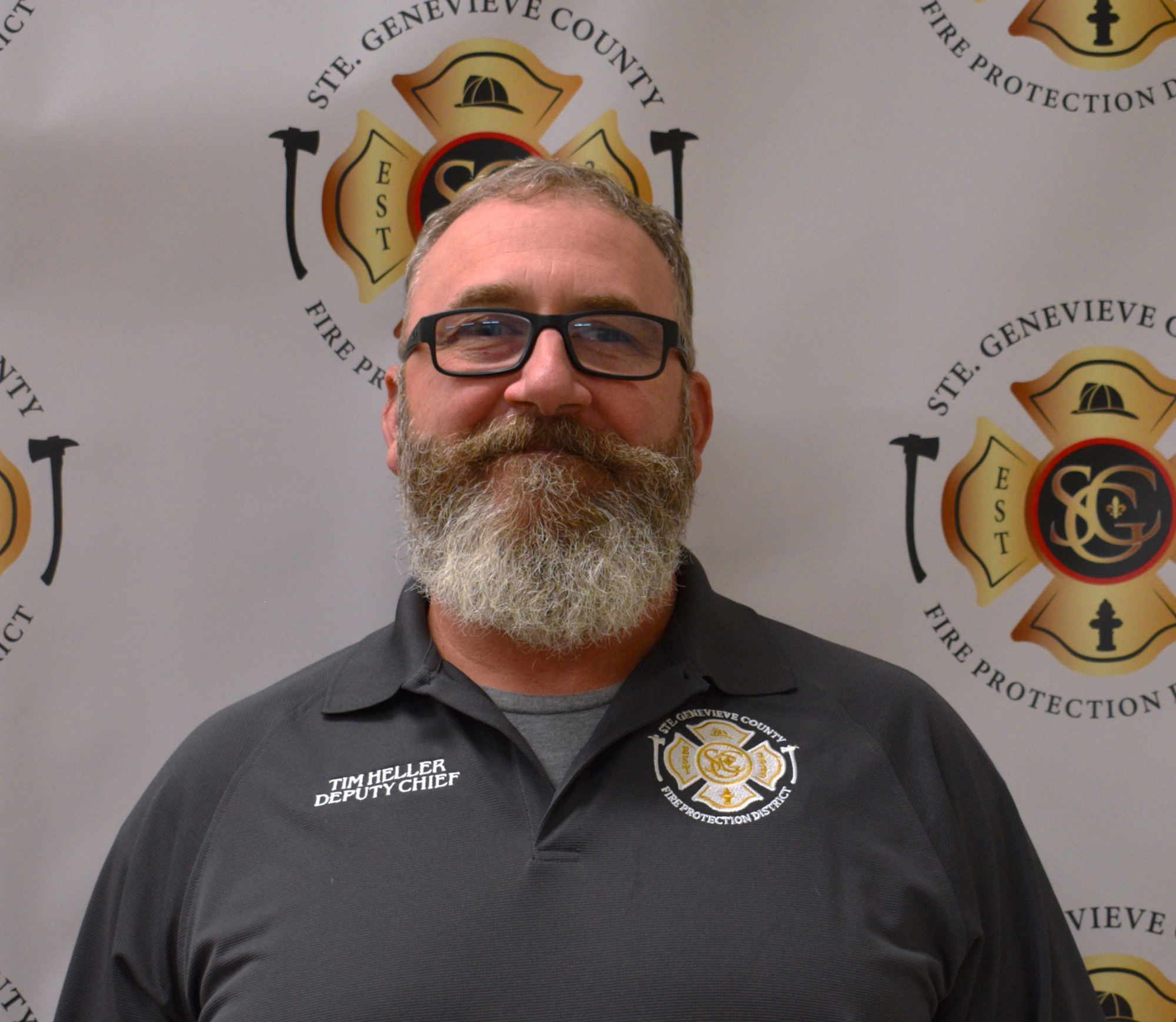 A man with a beard wearing a dark shirt stands in front of a backdrop featuring a fire protection district logo.