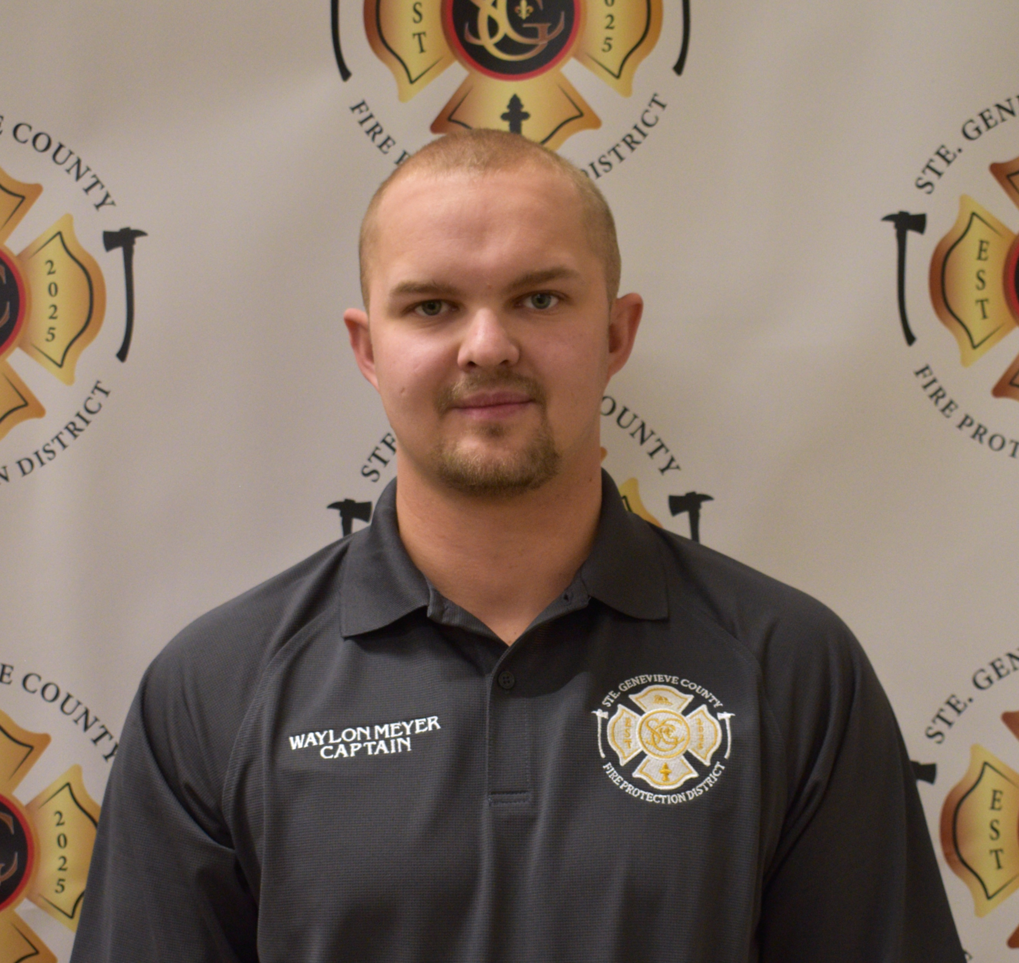 A man in a uniform with "Waylon Meyer Captain" printed on it, standing in front of a backdrop featuring a fire department logo.