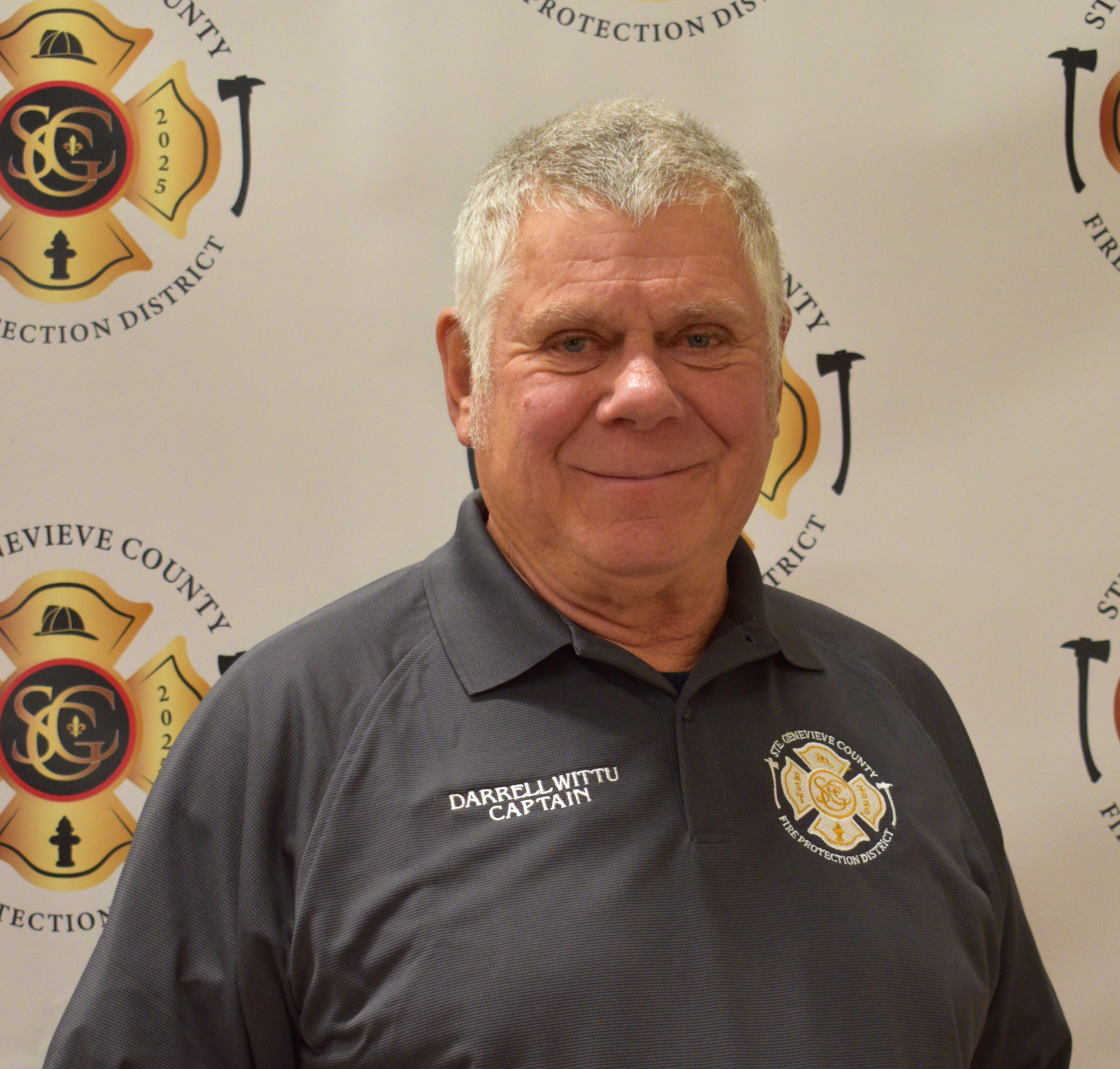 A smiling man in a captain's shirt poses in front of a fire protection district backdrop.