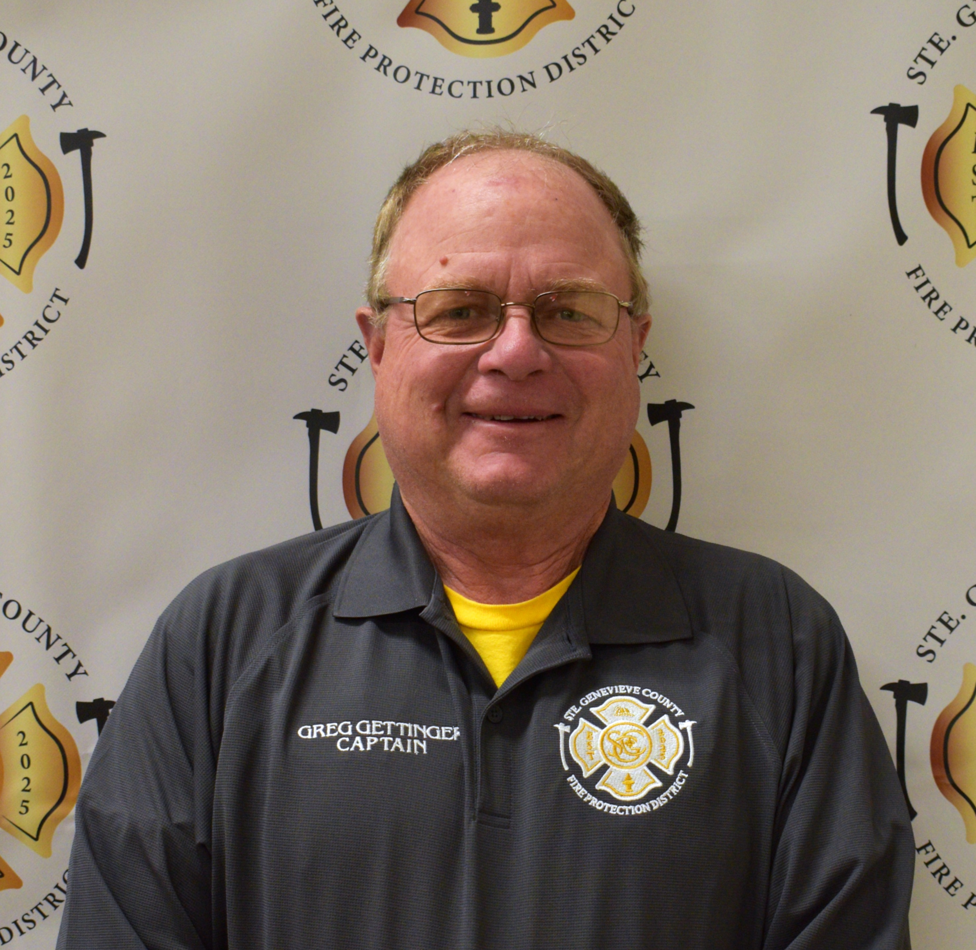 An official portrait of a man in a fire department uniform, smiling against a backdrop featuring fire protection insignia.