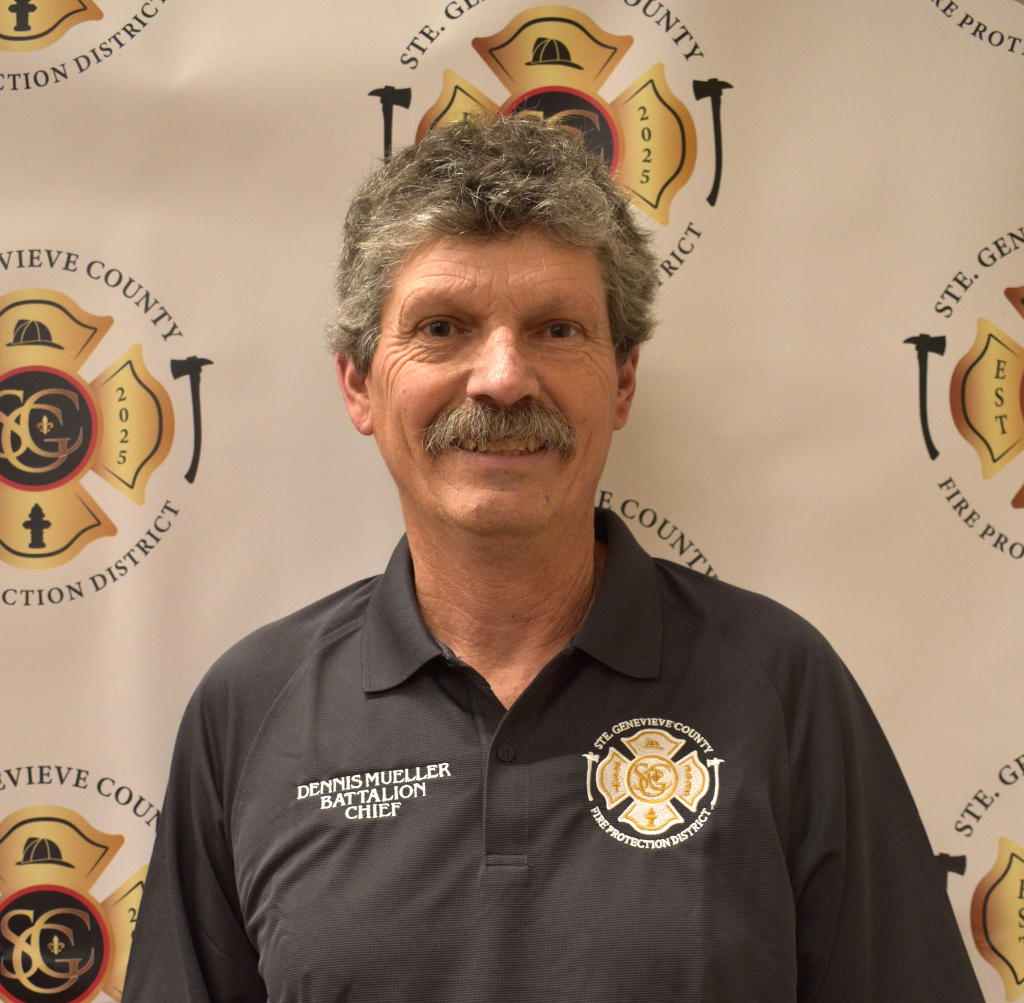 A man named Dennis Mueller, identified as Battalion Chief, poses in front of a fire protection district backdrop.