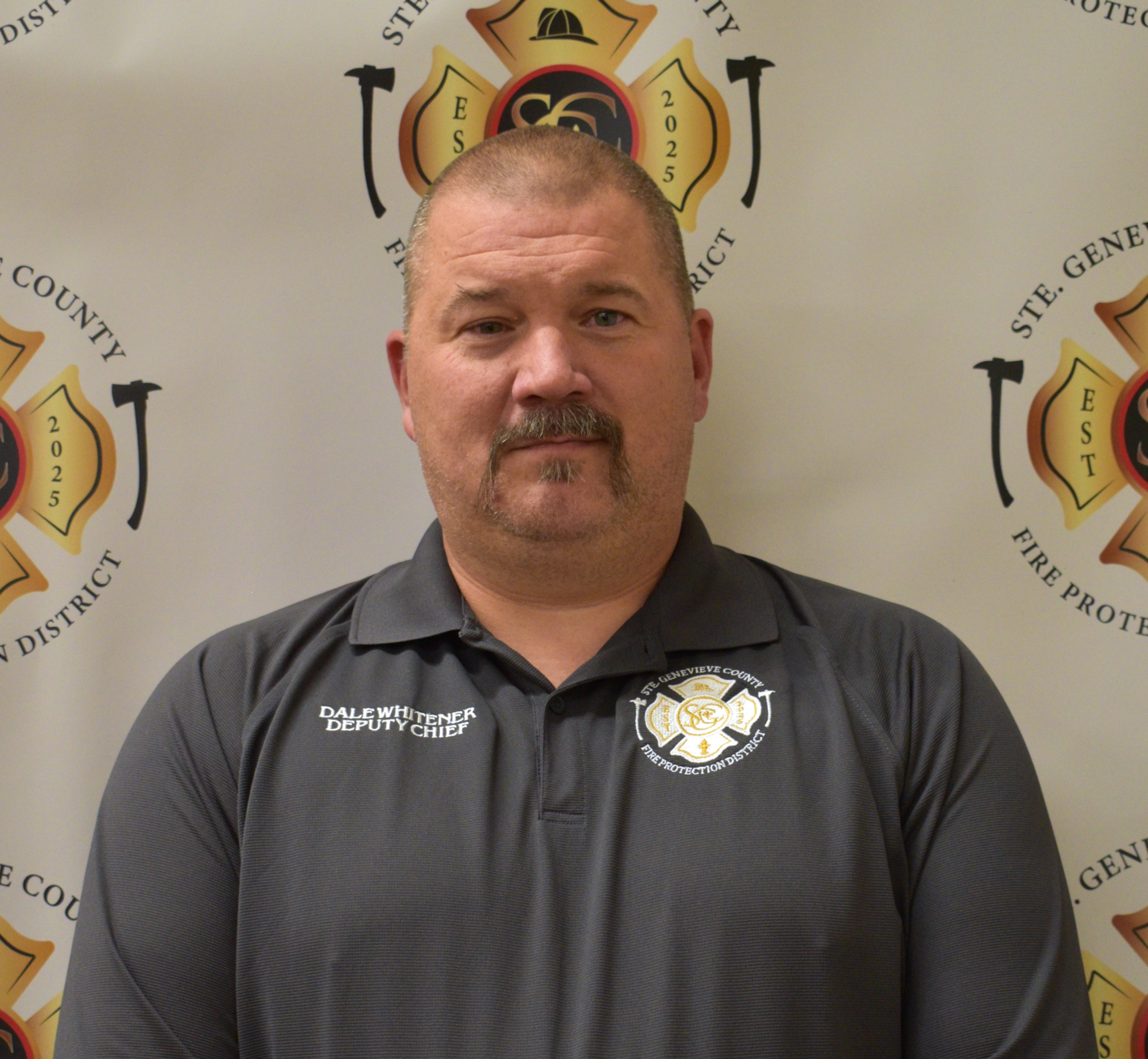 A man poses in a uniform with "Deputy Chief" and fire department logos in the background.