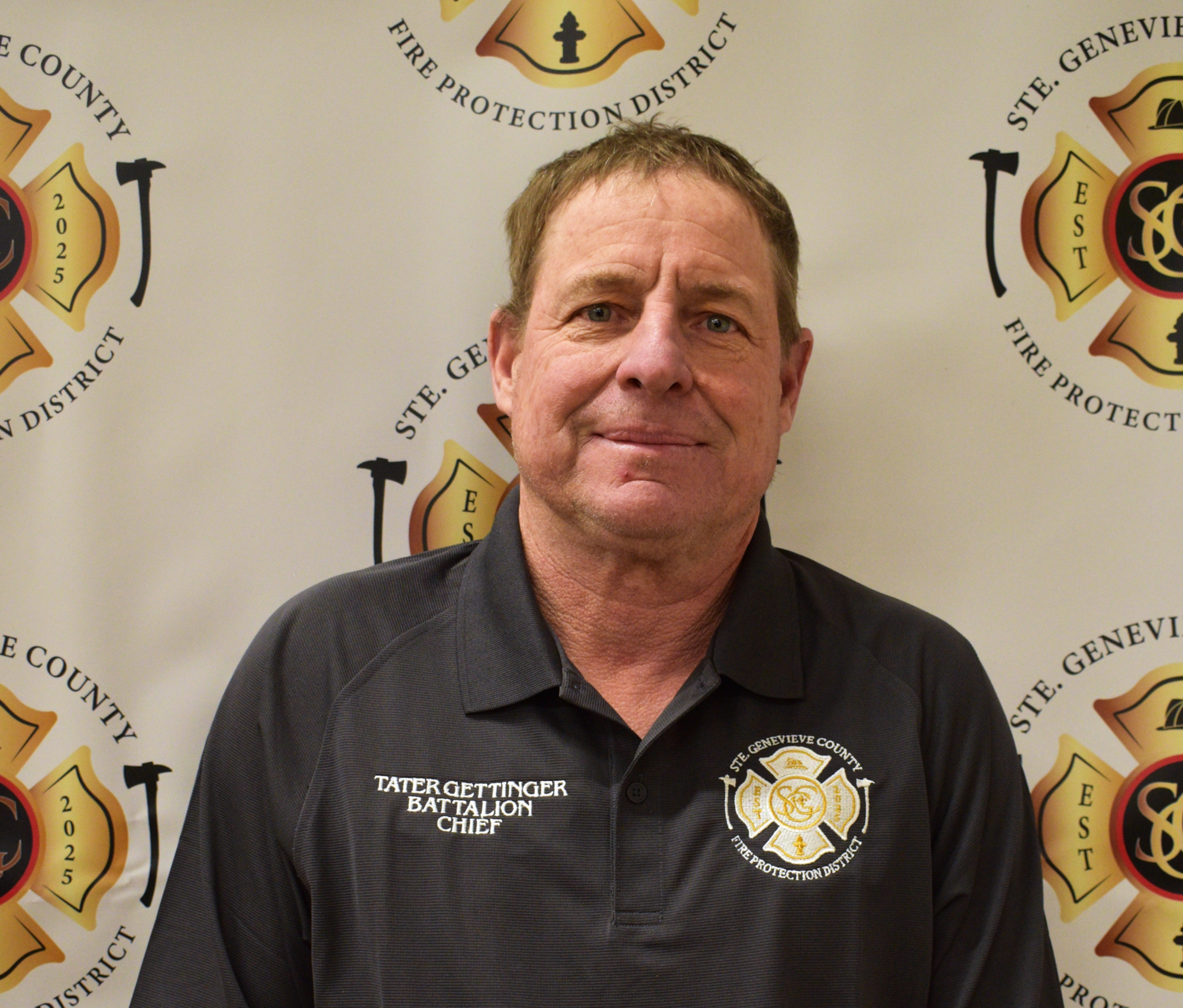 A man in a black shirt with a fire department logo, identified as a Battalion Chief, poses for a photo against a backdrop.