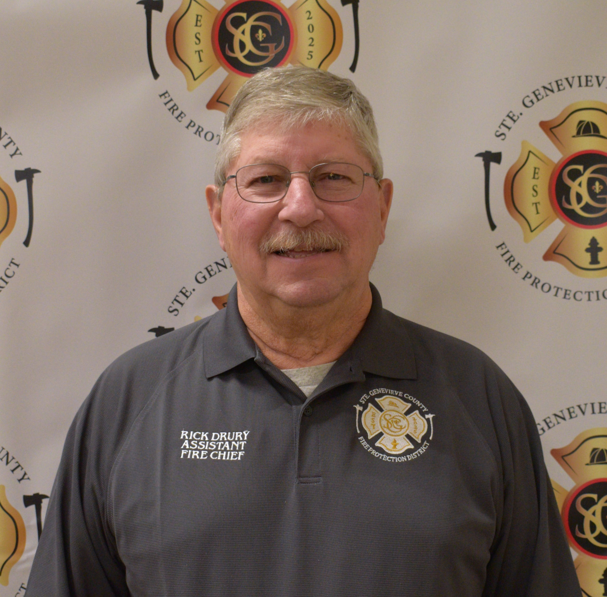 The image shows a smiling man in a gray polo shirt with "Assistant Fire Chief" and "Rick Drury" written on it, against a fire department backdrop.