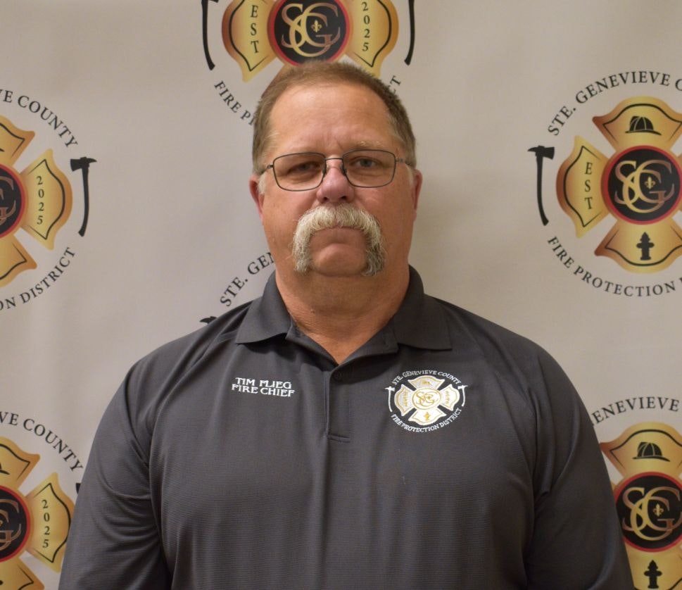 A man with glasses and a mustache wearing a fire chief uniform stands in front of a fire protection district backdrop.