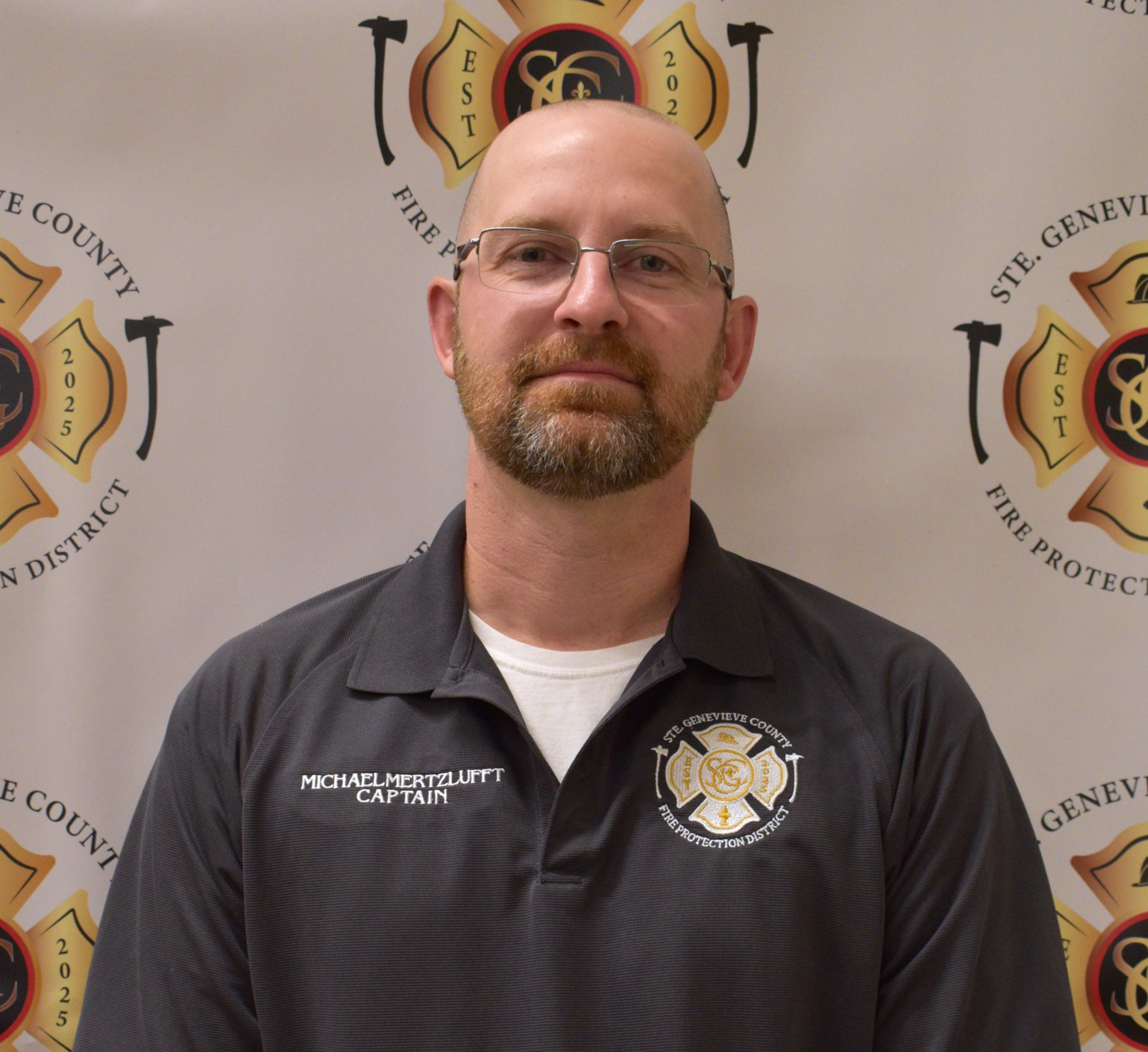 A man in a fire department shirt poses for a photo in front of a fire protection district background.