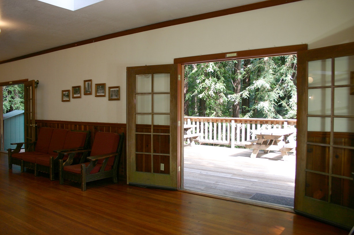 Indoor room with wooden floors and furniture, framed pictures on the wall, leading to an outdoor wooden deck surrounded by trees.