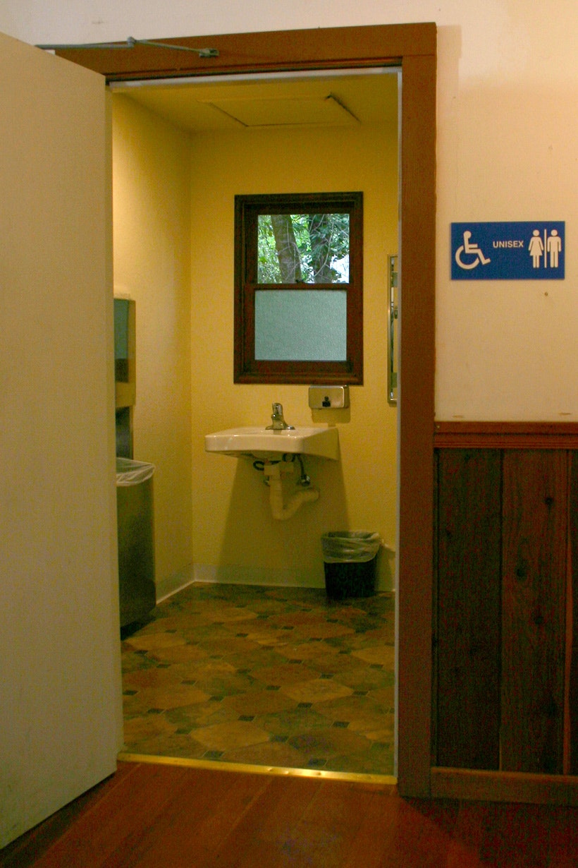Open door to a unisex bathroom with sink, towel dispenser, and trash bin visible. A sign indicates accessibility.