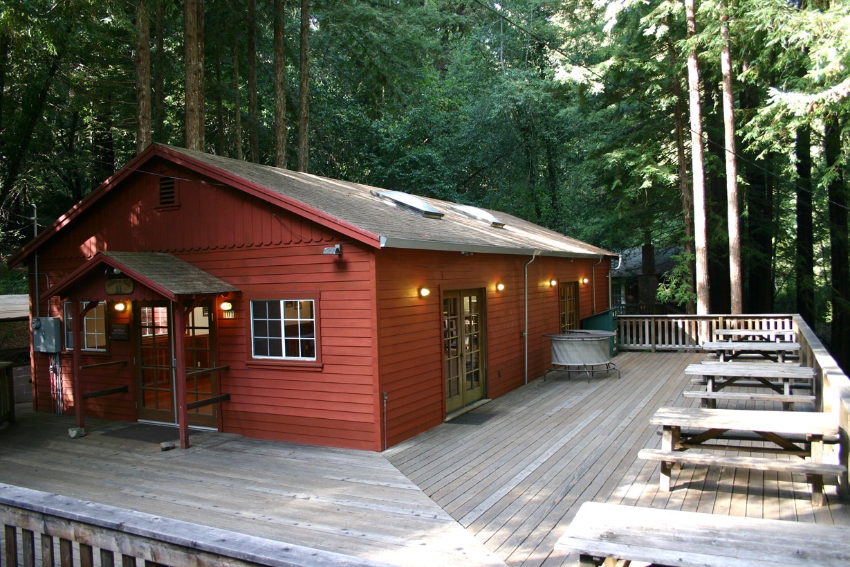 A red wooden cabin with a deck surrounded by tall trees.