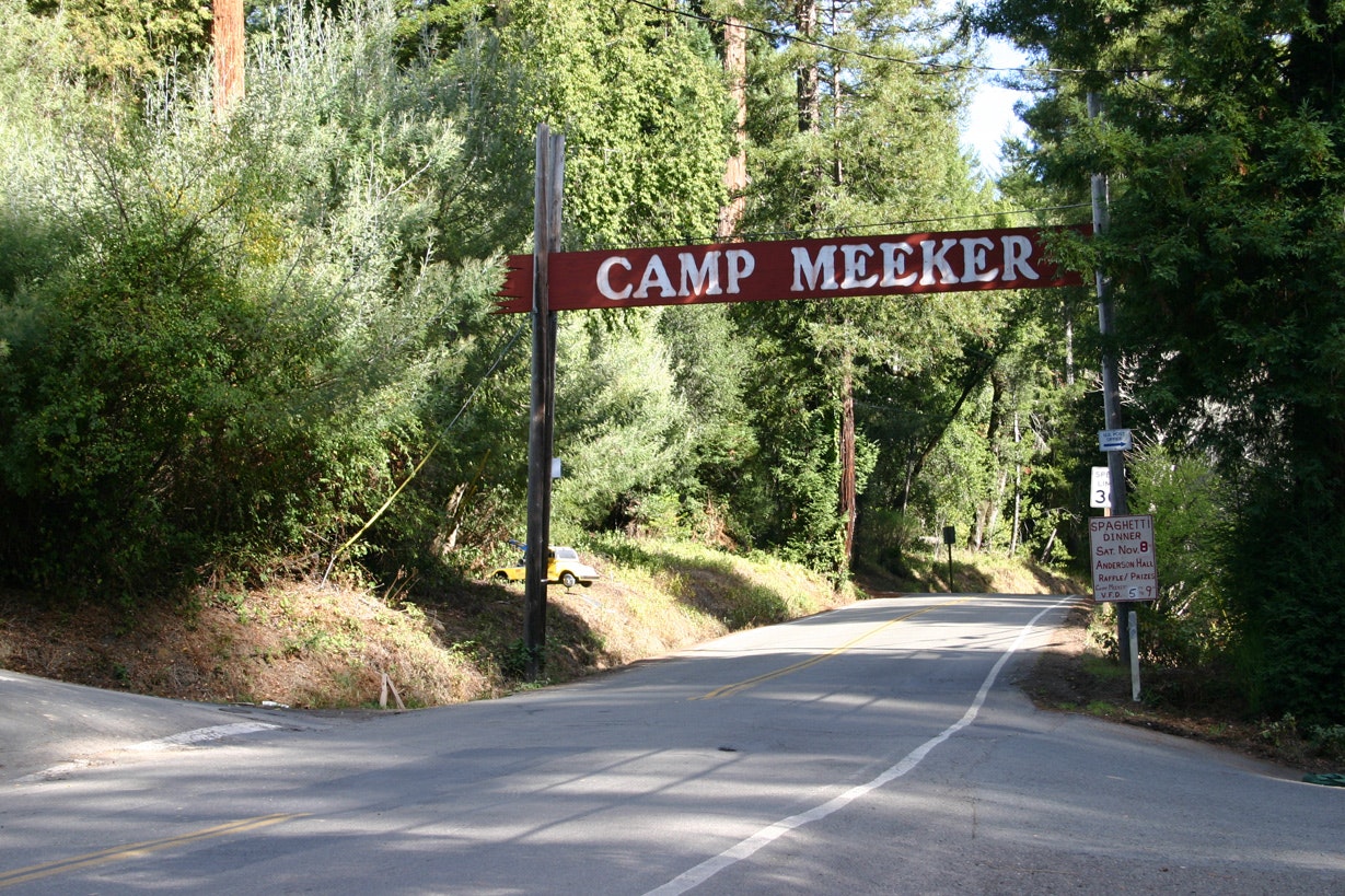 A road sign that reads "CAMP MEEKER" hanging across a road amidst a wooded area.