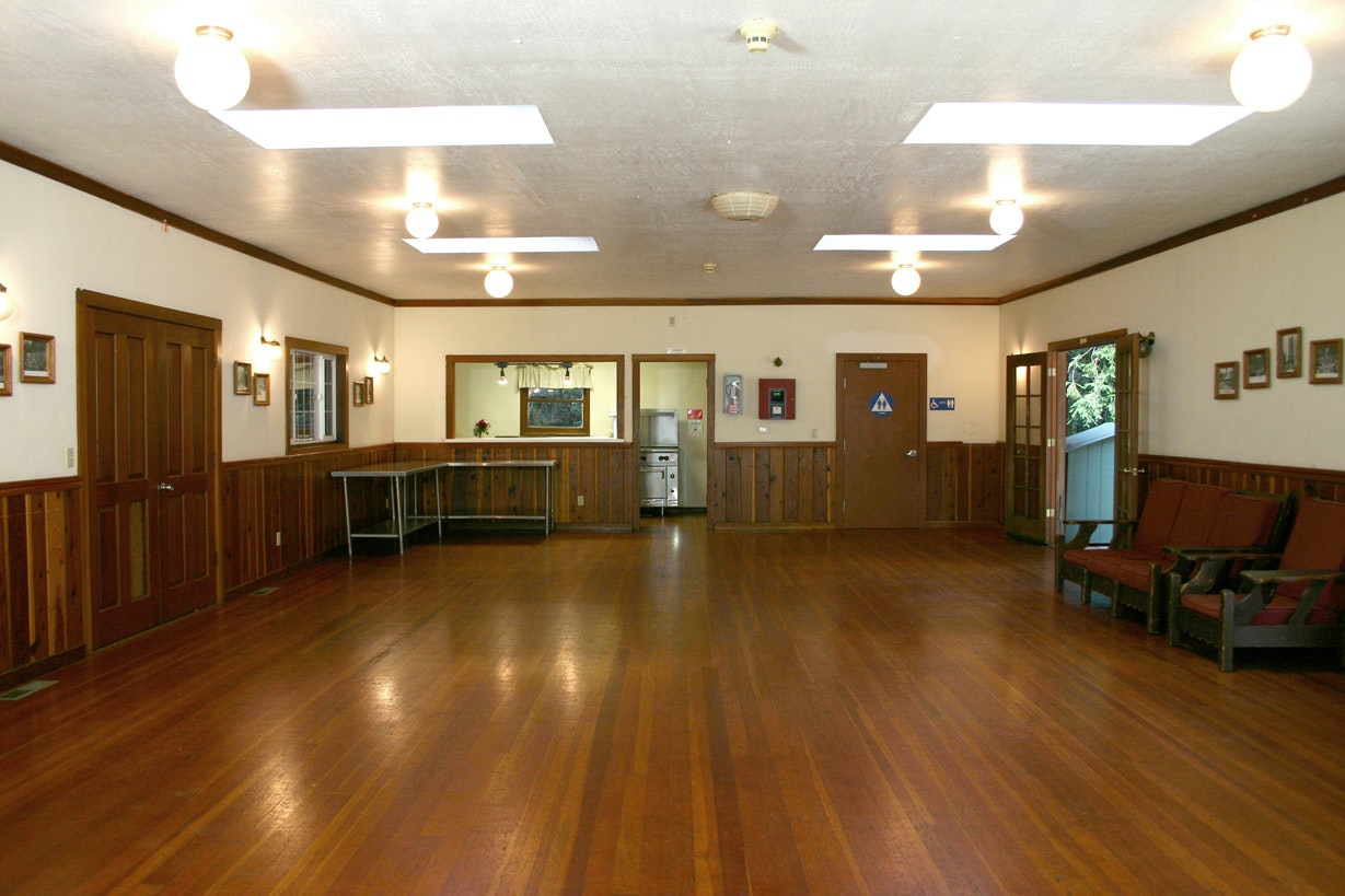 Empty hall with wooden floors, benches, doors, framed pictures, and fluorescent lights.