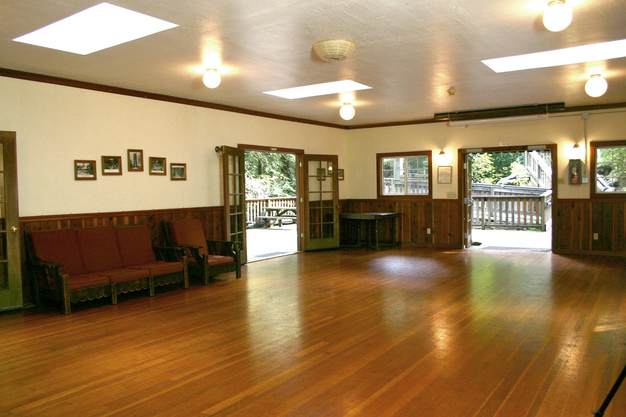 A well-lit, empty room with wooden floors, benches, framed pictures, and open doors leading to a deck.