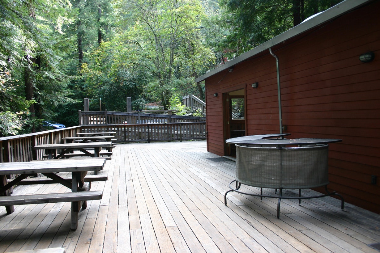 A wooden deck with a fire pit, picnic tables, and forest surrounding a red-painted building.