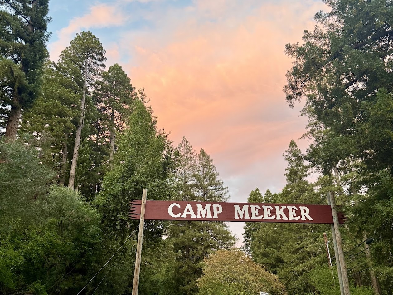 A wooden sign reads "Camp Meeker" surrounded by tall trees and a colorful sky at sunset.