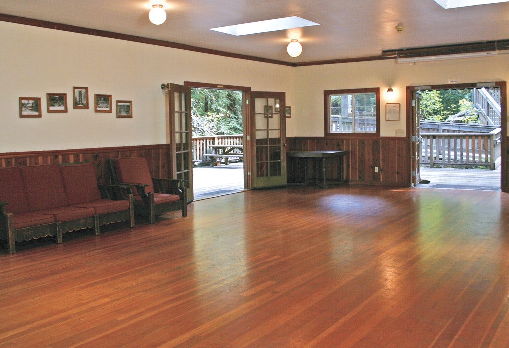Empty wooden-floored room with benches, opened doors to a deck, framed pictures on walls, and bright lighting.