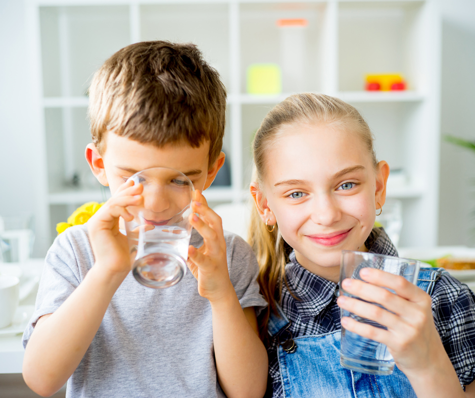 Two children smiling, holding and drinking glasses of water indoors.