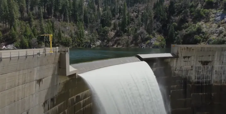 A dam releasing water into a reservoir, surrounded by lush forests and rocky terrain on a sunny day.