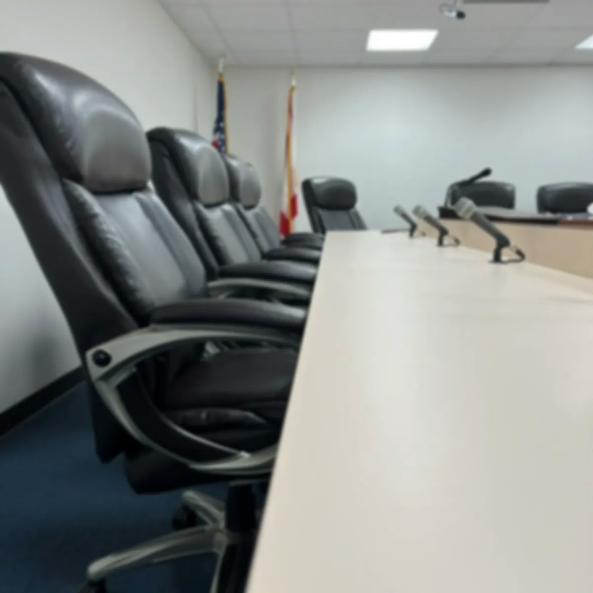 A blurred image of a conference room with a long table and several black office chairs. Flags are in the background.
