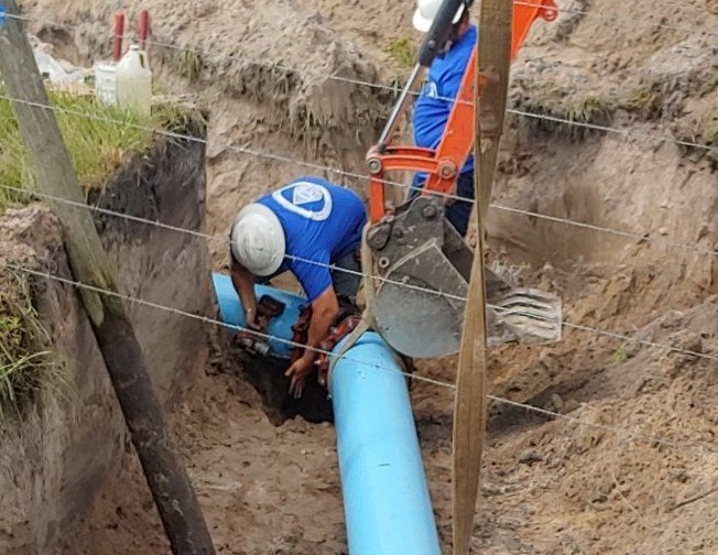 Workers in a trench are installing a large blue pipe, assisted by an excavator.
