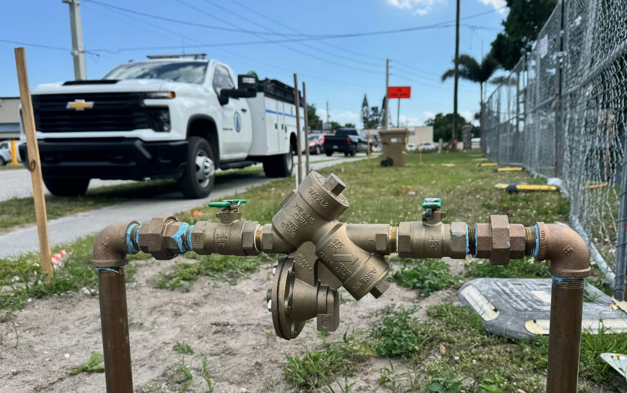 A brass backflow preventer on an outdoor pipe; a utility truck is in the background on a street.
