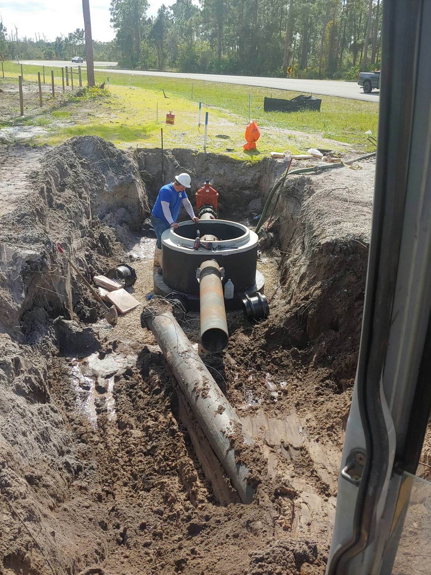 A worker inspects a large pipe in a dug trench near a road, surrounded by dirt and construction equipment.