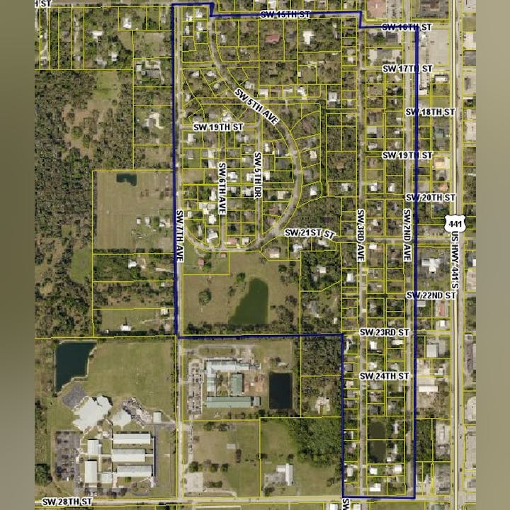 Aerial map showing roads, plots, and a residential area outlined in blue lines, with nearby greenery and buildings.