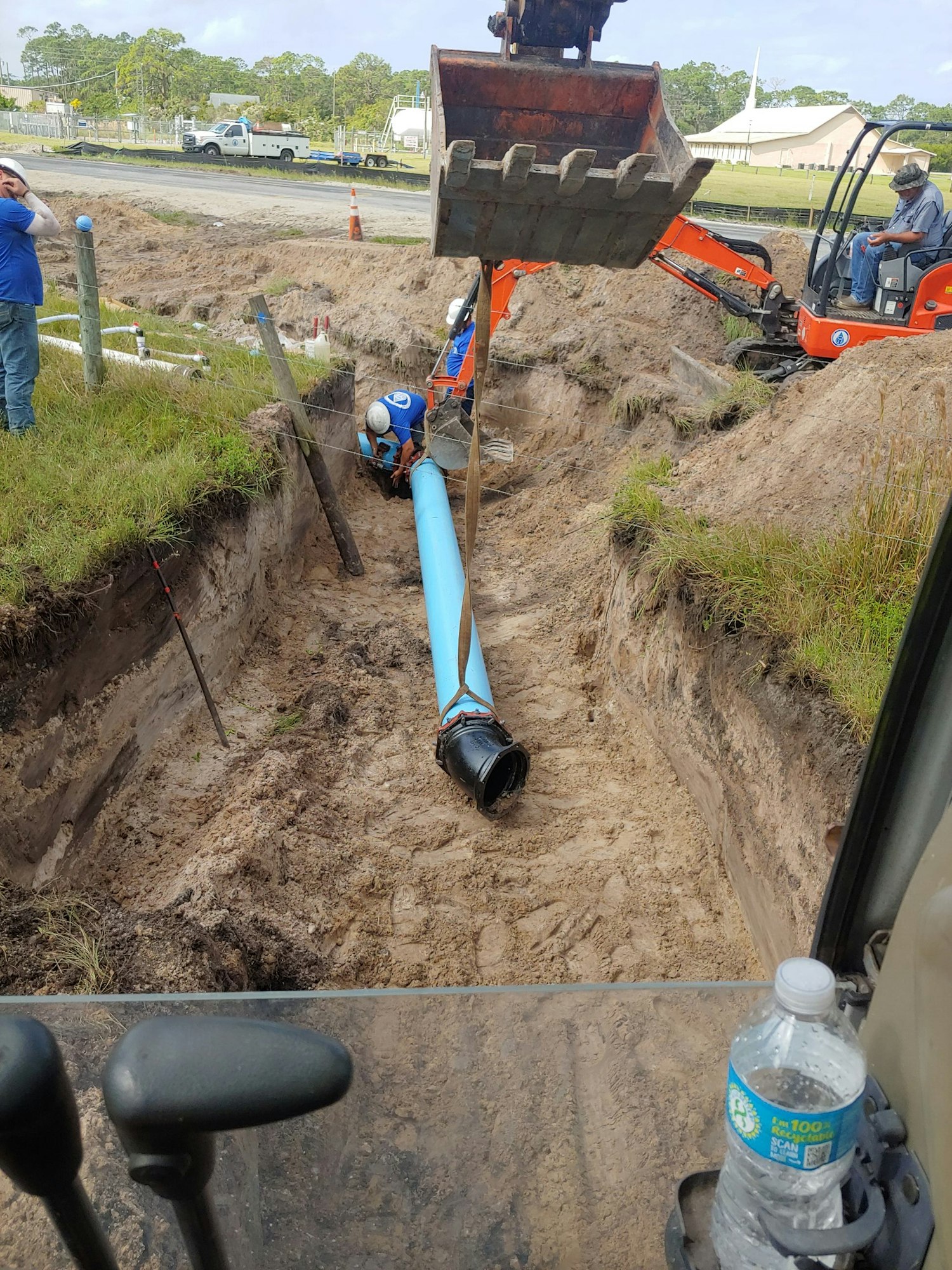 Construction site with workers installing a large blue pipe using an excavator in a trench.
