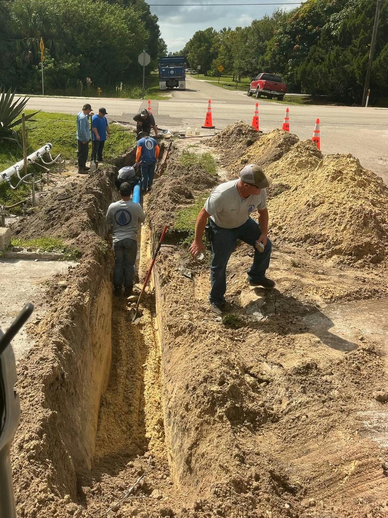 Workers digging a trench along a road, with traffic cones and a blue pipeline visible.