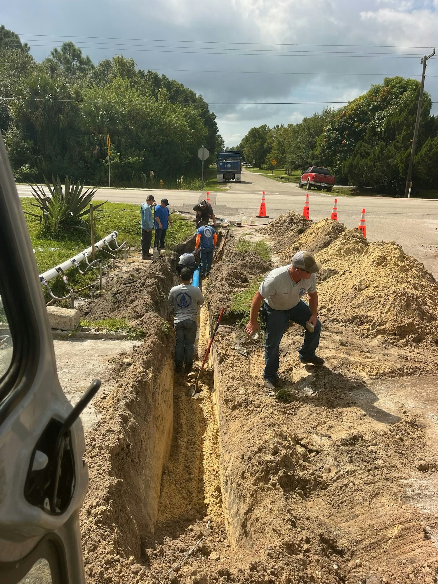 Workers digging a trench along a road, with traffic cones and a blue pipeline visible.