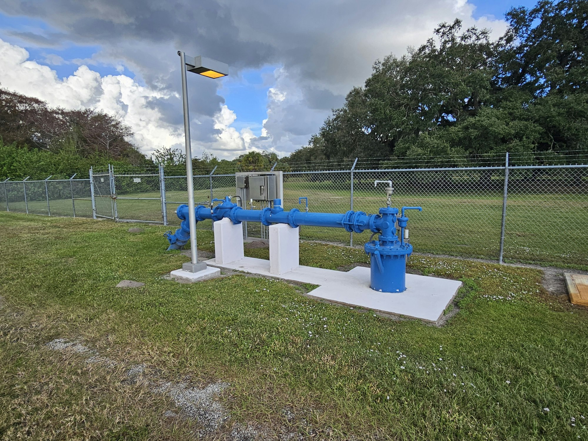 A blue pipeline structure with a light pole and fenced grassy area in the background.